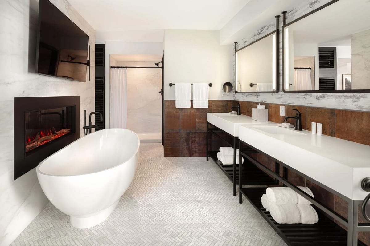 A hotel guest bathroom with a freestanding tub, linear fireplace and twin vanities
