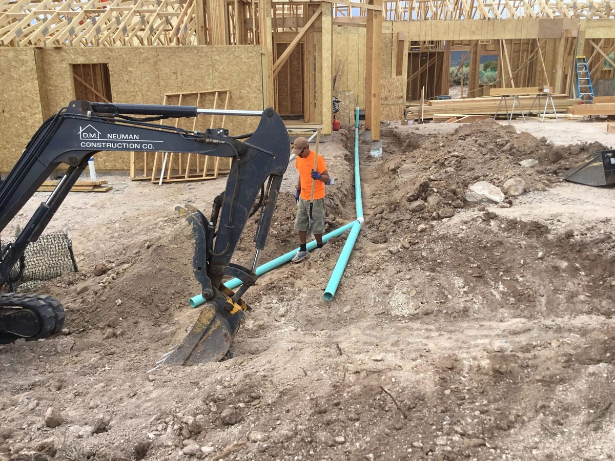 A mini excavator setting utility runs beside a house under frame